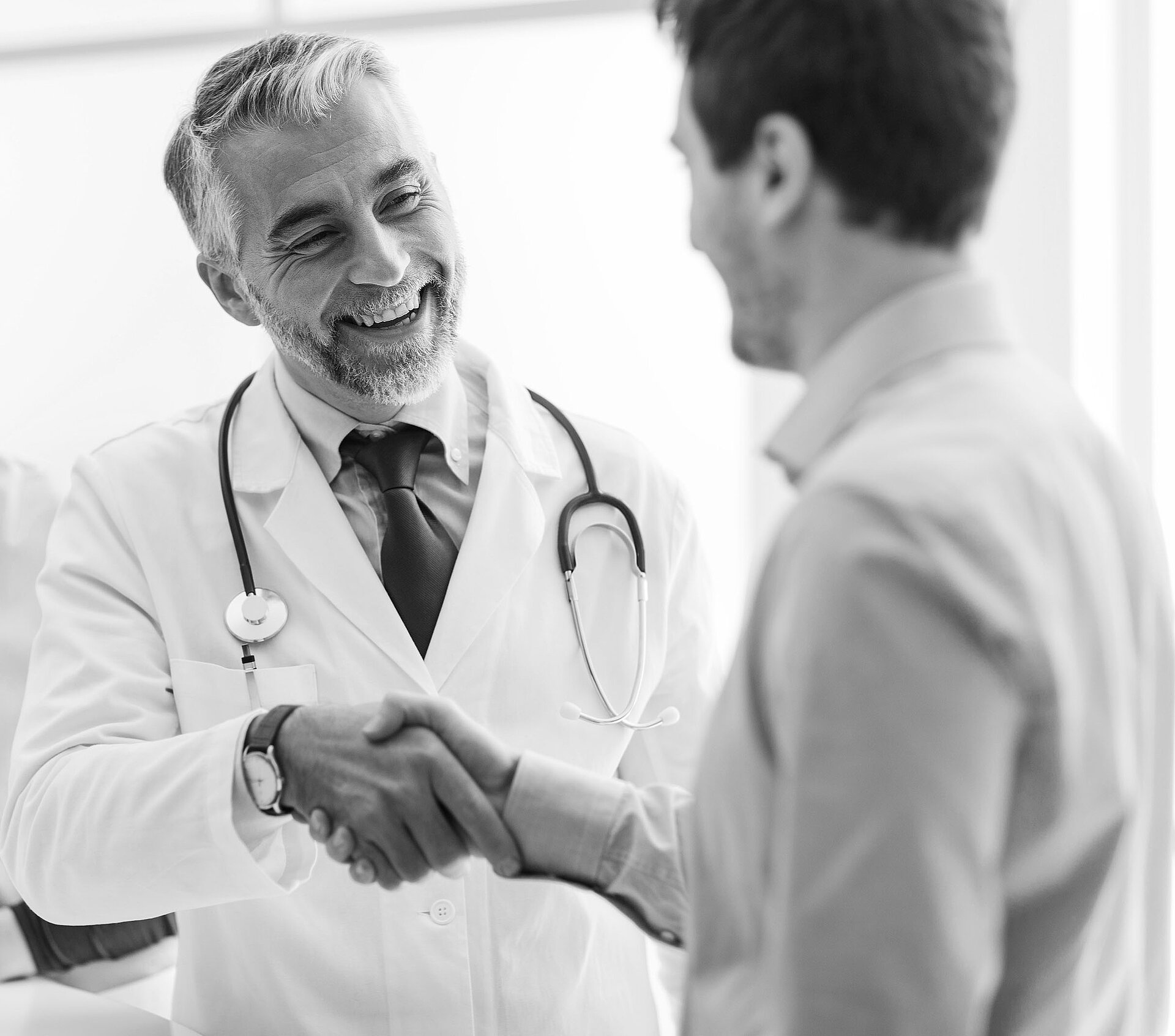 Black and white image of a middle-aged male doctor in a white coat and tie smiling at a male patient